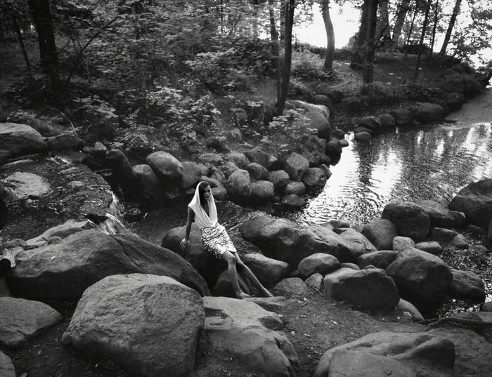 Black and white fashion editorial photograph of a model in a hooded top and patterned skirt sitting on river boulders in a forest.