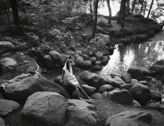 Black and white fashion editorial photograph of a model in a hooded top and patterned skirt sitting on river boulders in a forest.