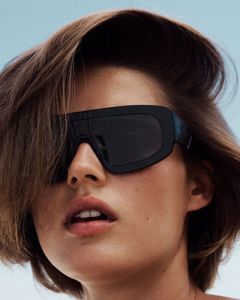 Close-up of a woman with short brown hair wearing large black shield sunglasses against an open sky.