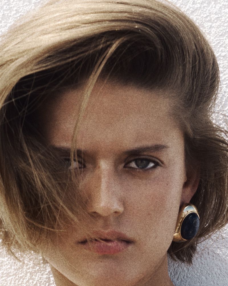 Close-up portrait of Bonnie Panasenko with side-swept hair wearing a chunky gold and dark stone earring against a white wall.