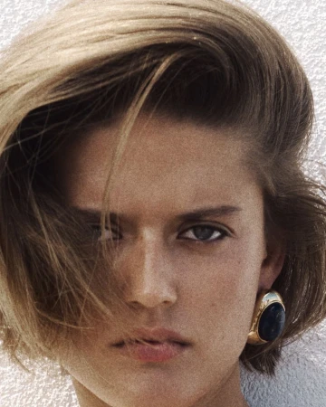 Close-up portrait of Bonnie Panasenko with side-swept hair wearing a chunky gold and dark stone earring against a white wall.