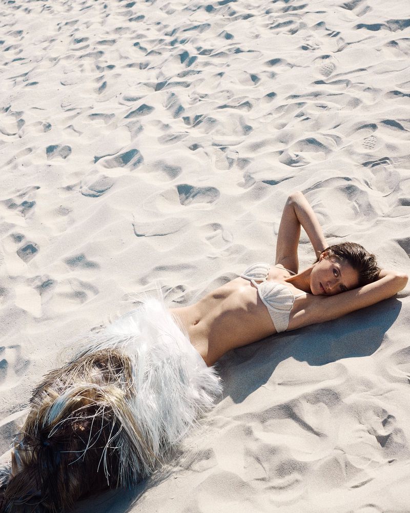 Model Bonnie Panasenko lying on white sand wearing a seashell bikini top and textured feathered skirt for Vogue Turkey.