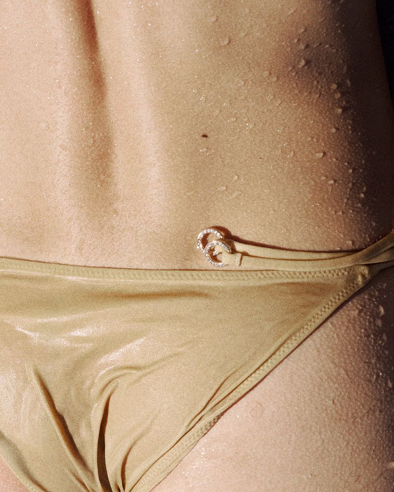 Close-up detail of wet skin with water droplets and a metallic gold bikini bottom featuring crystal ring hardware.