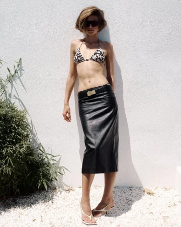 Model Bonnie Panasenko posing in a zebra print bikini top and black leather midi skirt against a white wall in direct sunlight.
