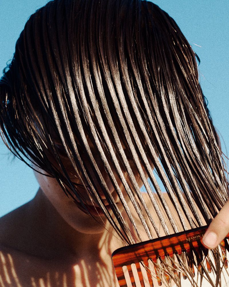 Close-up of wet dark hair combed over a model's face with a tortoiseshell wide-tooth comb against a clear blue sky.