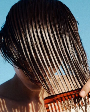 Close-up of wet dark hair combed over a model's face with a tortoiseshell wide-tooth comb against a clear blue sky.