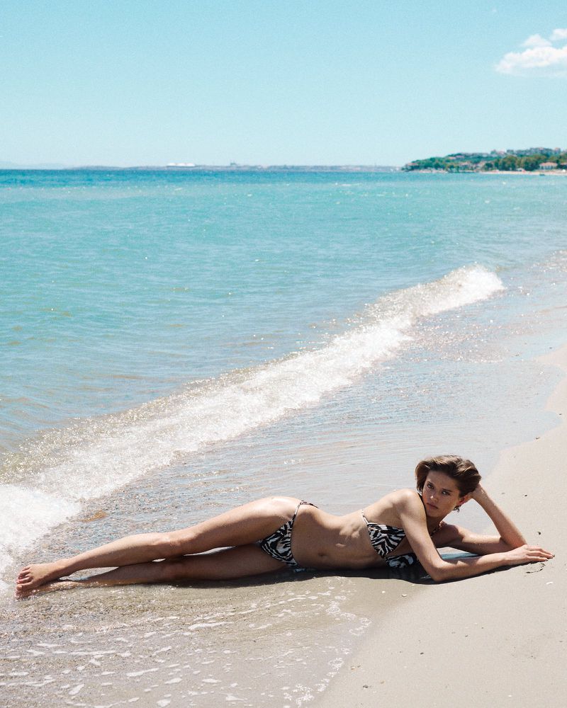 Model Bonnie Panasenko reclining on a sandy beach in a black and white patterned bikini by the ocean shore.