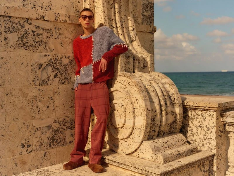 Fashionable adult male model wears red and gray patchwork sweater and plaid pants on a sunny stone terrace with ocean view backdrop.