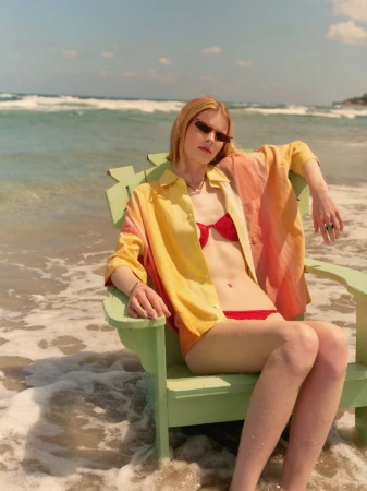 Blonde woman in red bikini and tie-dye shirt sits on a green chair on a beach with ocean waves in the background on a sunny day.