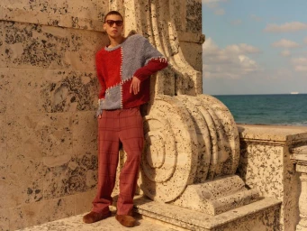 Model Jacoby Sorapuru wearing a red and grey patchwork knit sweater and plaid trousers leaning against a stone structure by the sea.