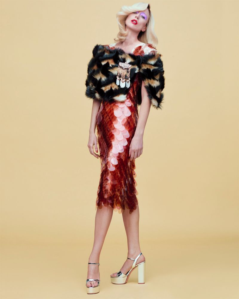 Model Raquel Zimmermann poses in a tiered red scale dress and patterned fur capelet against a pale yellow studio backdrop.