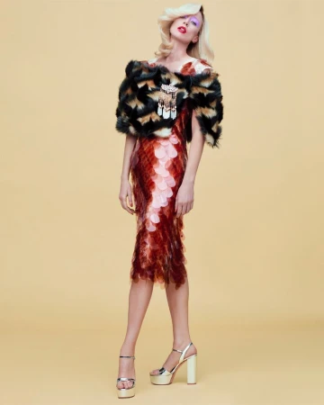 Model Raquel Zimmermann poses in a tiered red scale dress and patterned fur capelet against a pale yellow studio backdrop.