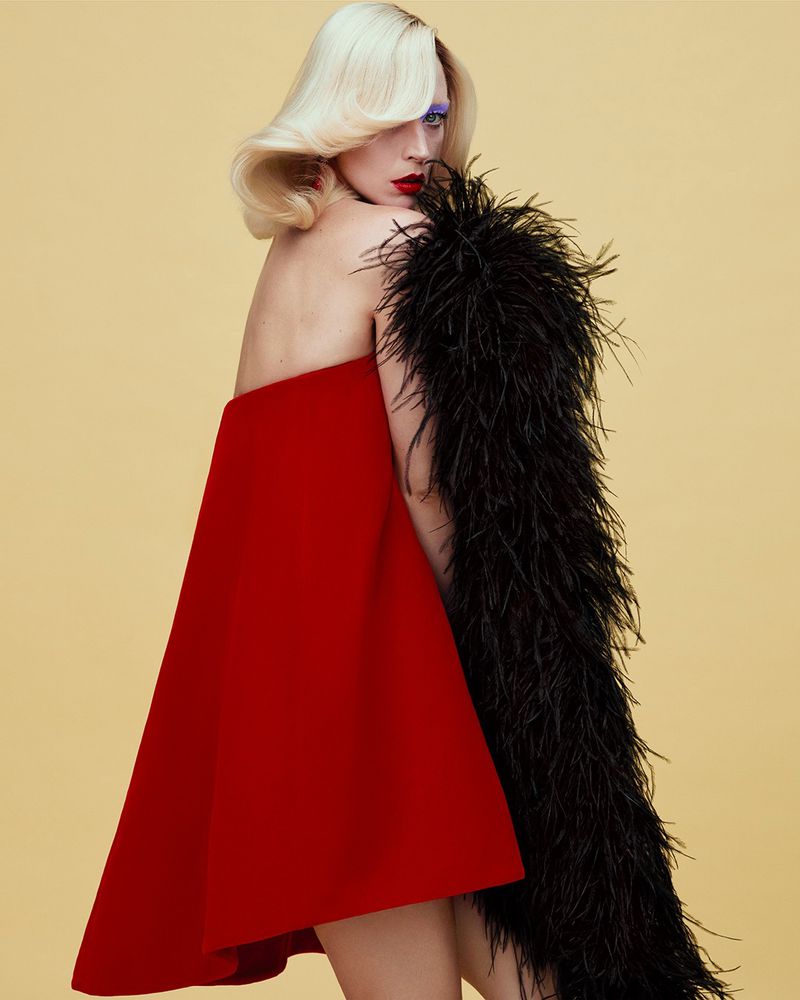 Model Raquel Zimmermann wearing a red trapeze mini dress and a black feather boa against a pale yellow backdrop.