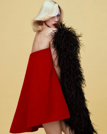 Model Raquel Zimmermann wearing a red trapeze mini dress and a black feather boa against a pale yellow backdrop.