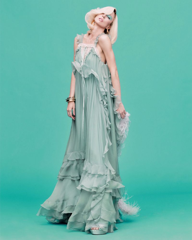 Model Raquel Zimmermann poses in a tiered ruffled sage green gown with gold bracelets against a solid turquoise backdrop.