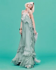 Model Raquel Zimmermann poses in a tiered ruffled sage green gown with gold bracelets against a solid turquoise backdrop.
