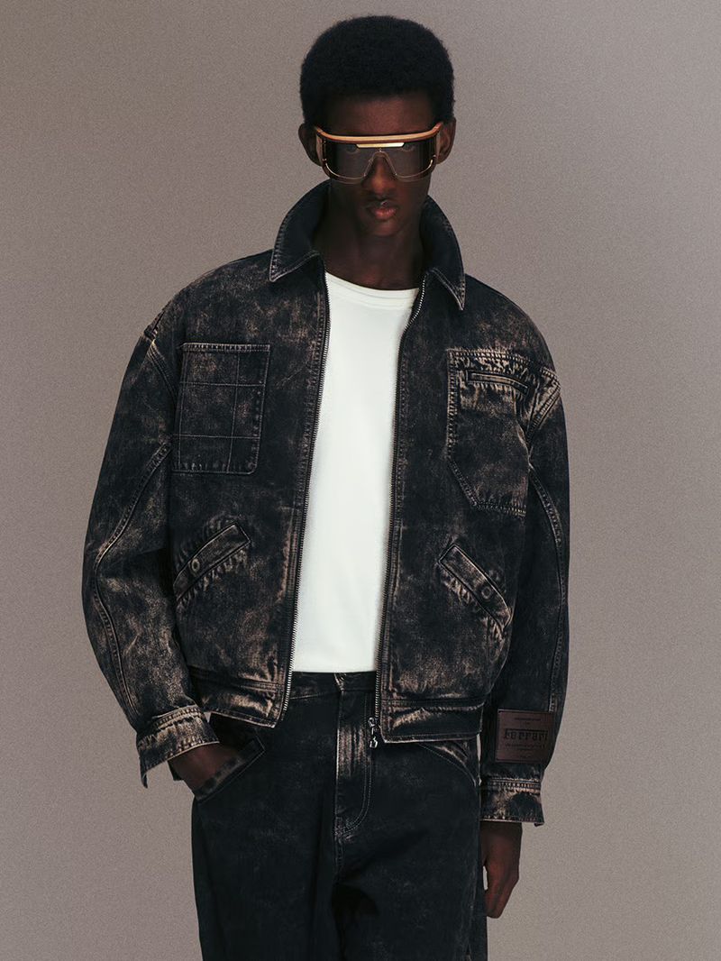 Model wearing a dark distressed denim jacket and tinted sunglasses against a neutral studio background