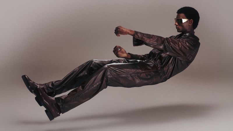 Model Yahne Ba floating in a driving pose wearing a dark glossy jumpsuit and visor sunglasses against a grey studio background.