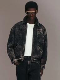 Model wearing a dark distressed denim jacket and tinted sunglasses against a neutral studio background