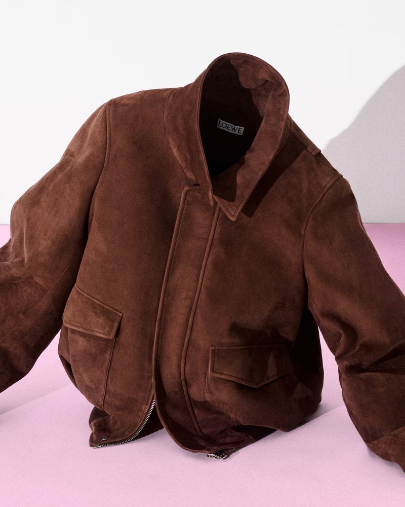 Brown suede bomber jacket with flap pockets positioned upright on a pale pink surface against a white backdrop.