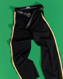 Black Loewe trousers with yellow and white side stripes and a black leather belt laid flat on a solid green background.