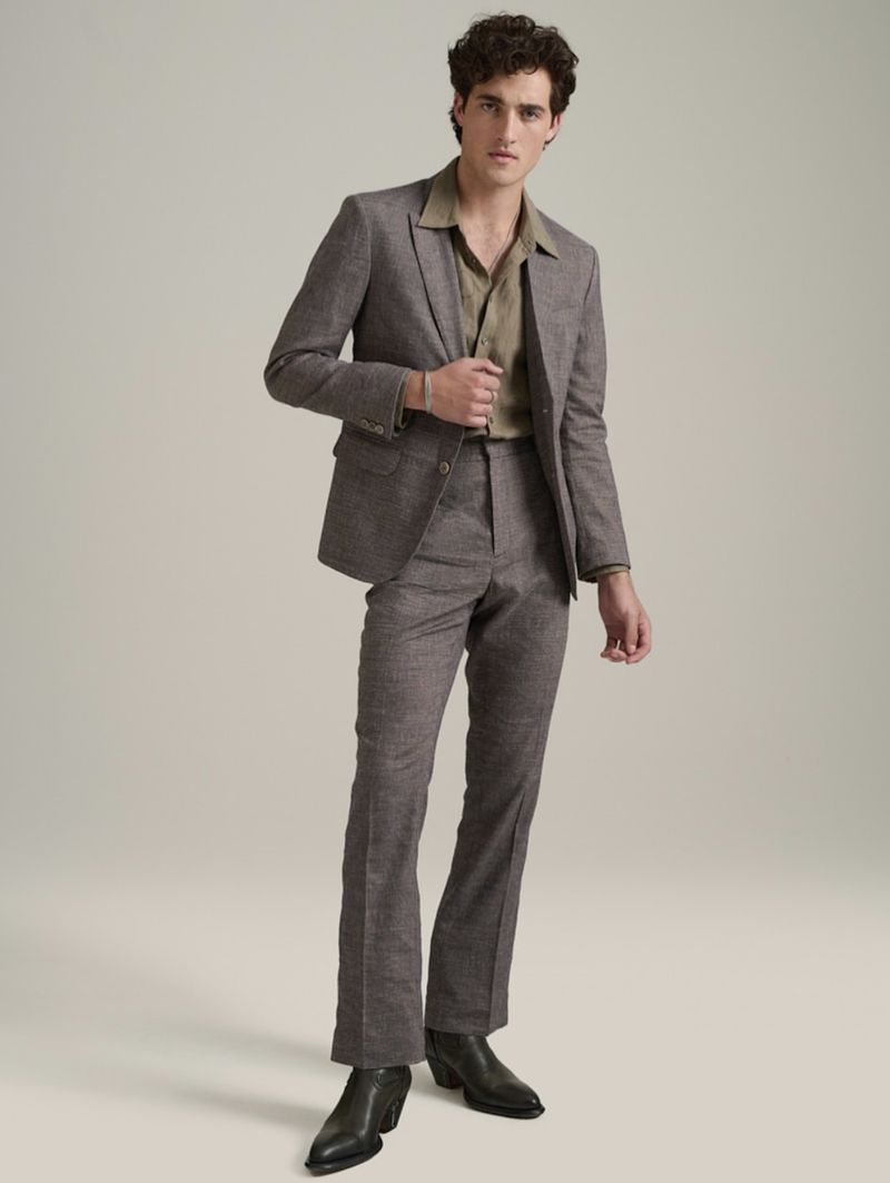 A man stands in a photo studio wearing a brown suit with an unbuttoned olive green shirt and black boots against a neutral background.