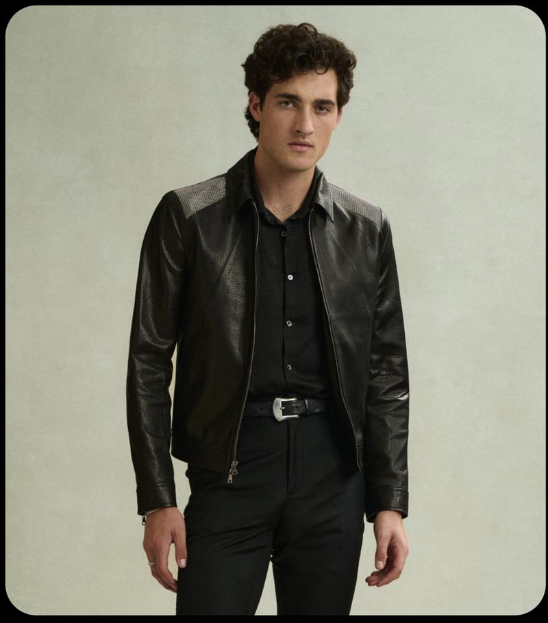 Portrait of a young adult male fashion model with curly brown hair wearing a black leather jacket and black shirt against a neutral background.