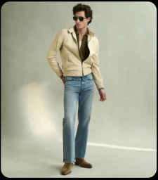 Man wearing sunglasses, cream jacket, green polo, and blue jeans standing against a neutral studio backdrop