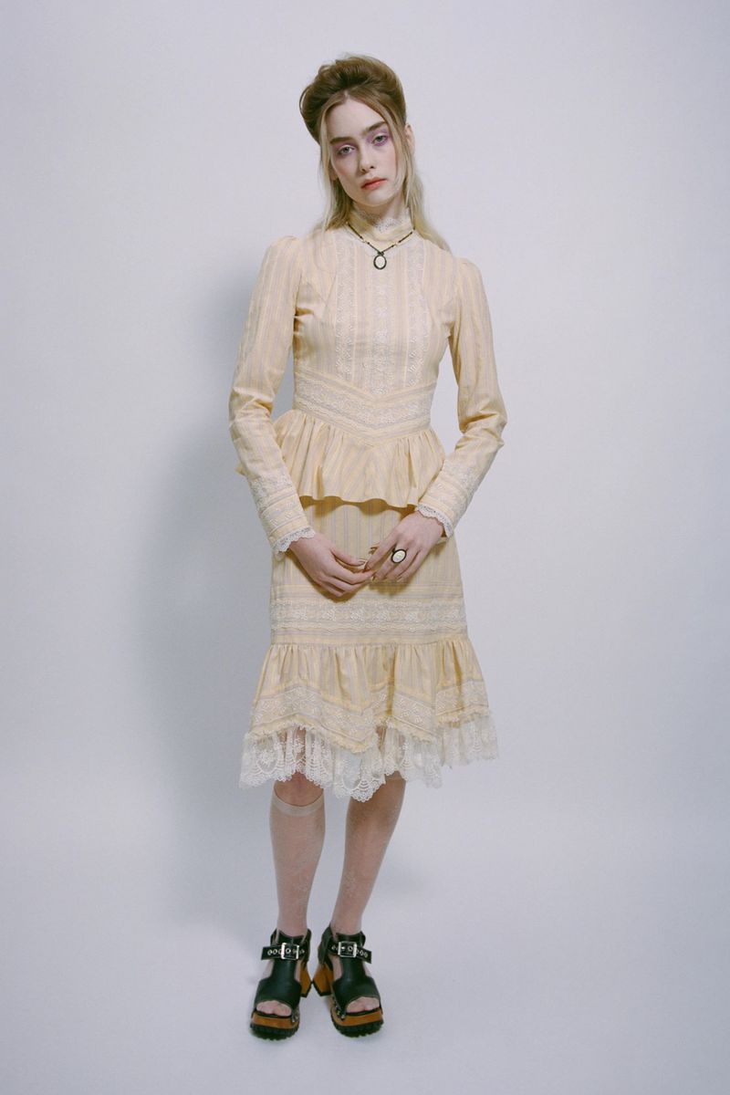 Full-length portrait of a blonde woman in an embroidered high-necked dress and platform sandals in a white studio setting. 