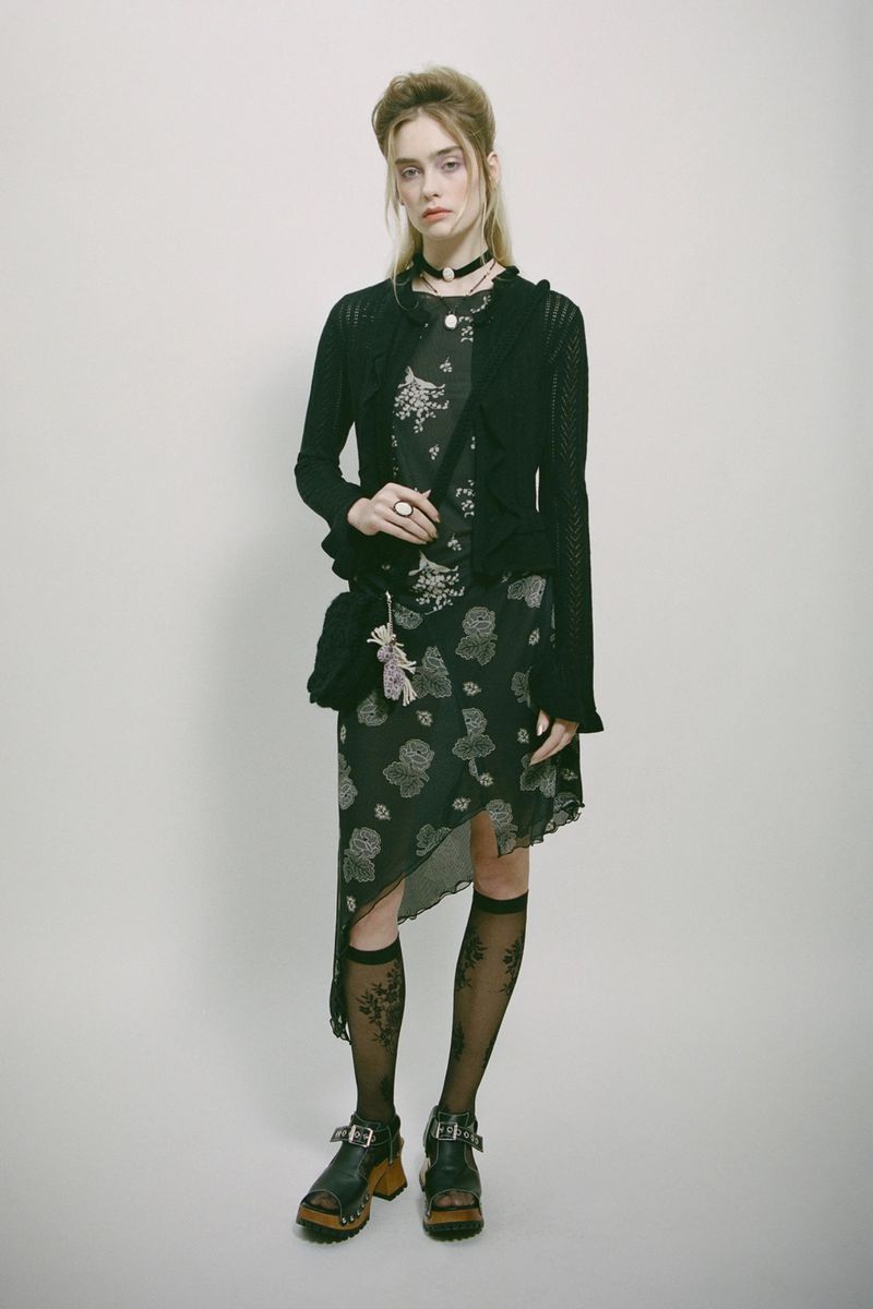A young woman poses in a black floral print dress with a black cardigan, decorative socks, and platform shoes against a white backdrop.