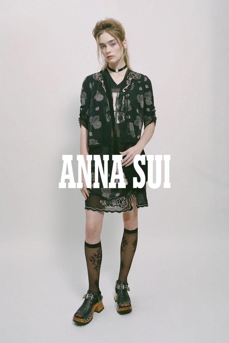 A slender fashion model poses in a patterned black jacket, sheer skirt, knee socks, and platform shoes against a plain background.