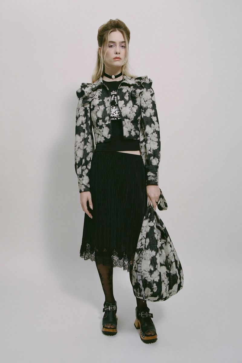 A fair-skinned young adult female poses in a studio wearing a floral print jacket and pleated skirt, holding a matching tote bag.