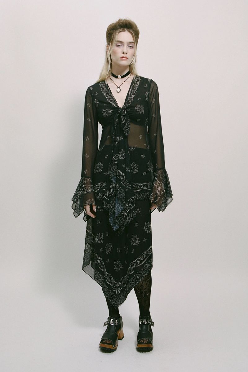 A young adult woman poses in a full-length shot, wearing a black patterned semi-sheer midi dress and black chunky platform shoes on a white backdrop.