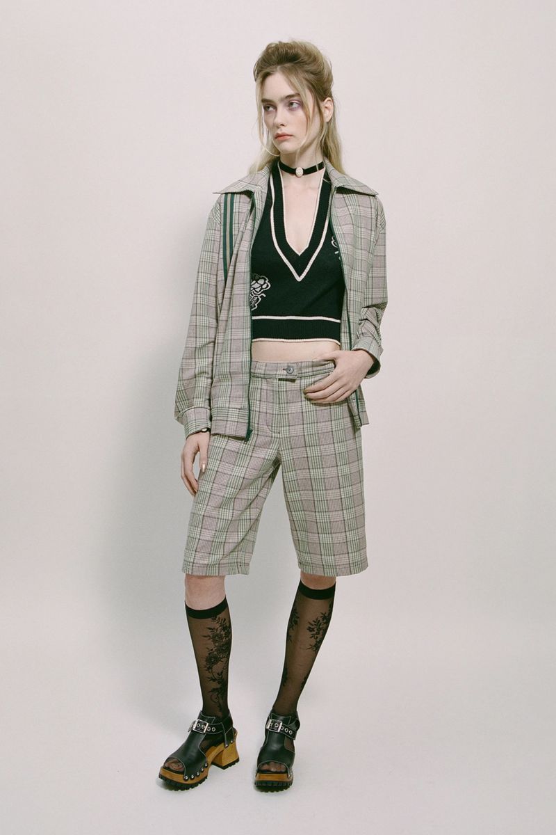 Full-shot of a slender, fair-skinned young adult woman in a plaid blazer and shorts, posed against a bright studio backdrop. She has semi-sheer patterned socks.