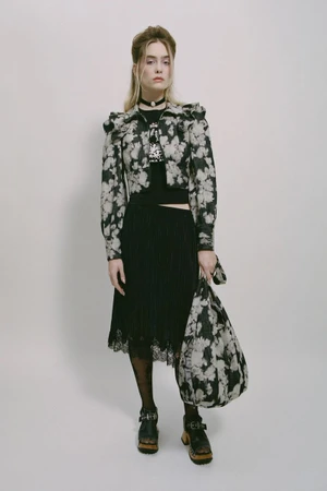 Model wearing a patterned floral jacket, black pleated skirt, and holding matching bag against a white seamless backdrop.