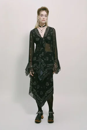 Model wearing a black patterned sheer dress with flared sleeves and buckled platform sandals against a plain grey backdrop.