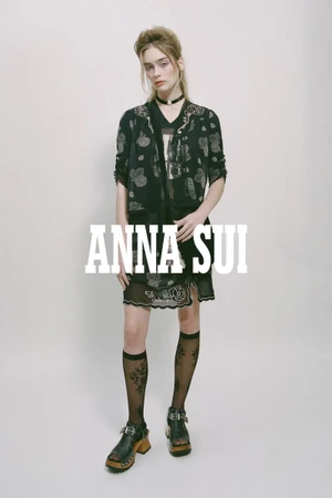 Model wearing a black patterned dress and stockings standing against a plain white background for Anna Sui