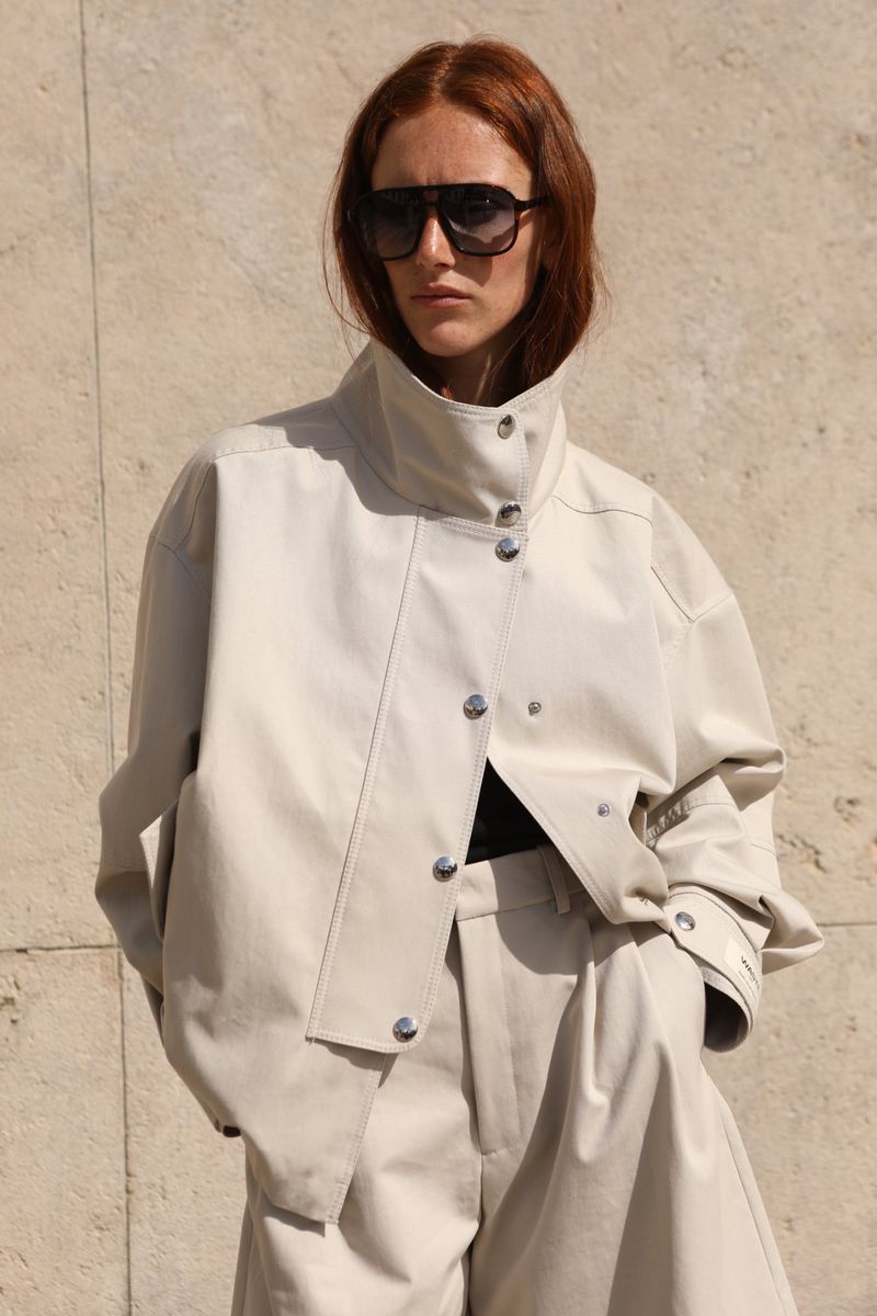Red-haired model wearing oversized cream jacket and sunglasses against stone wall