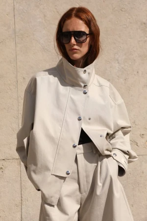 Red-haired model wearing oversized cream jacket and sunglasses against stone wall