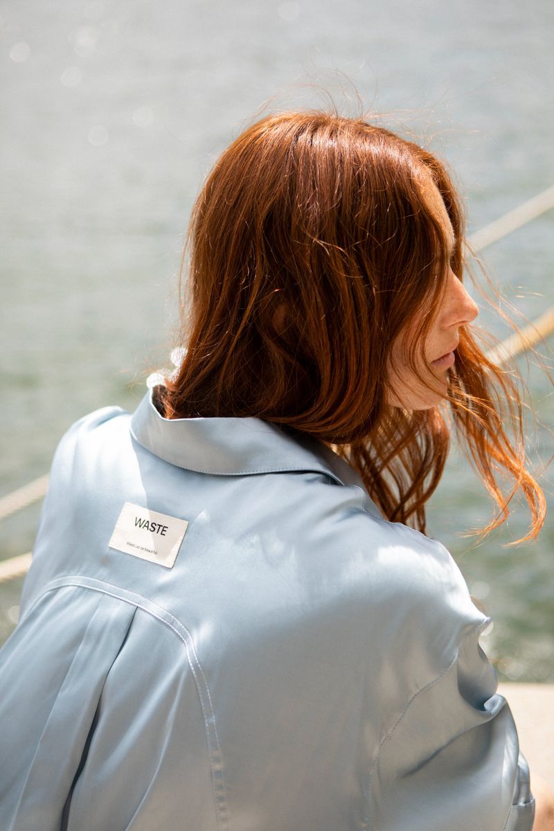 Woman with auburn hair seen from behind wearing a light blue satin shirt with a Waste tag on the upper back by water.