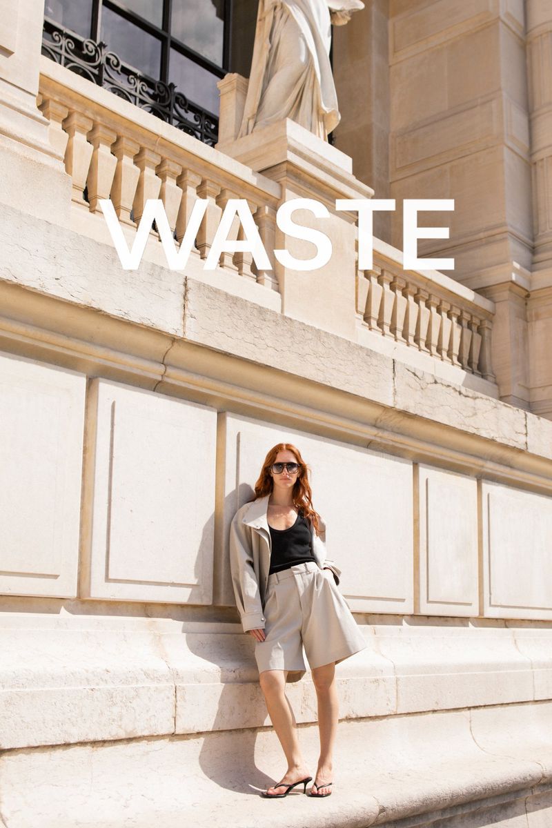 Model with red hair wearing sunglasses, cream jacket, black top, and beige shorts leaning against a stone building exterior