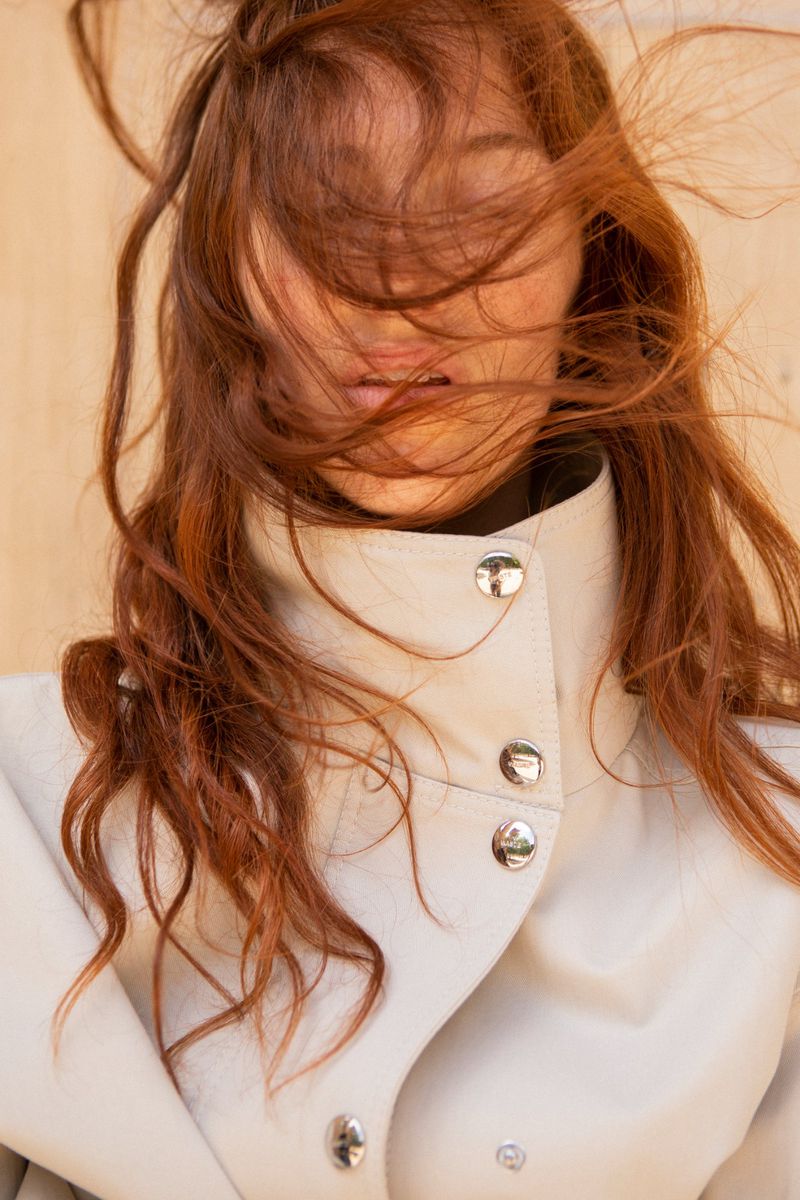Close-up of a woman with windblown auburn hair covering her face, wearing a high-collared off-white coat with silver buttons.