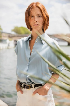 Redhead woman in a pale blue button-up shirt and white trousers standing by a riverbank with hands in pockets.
