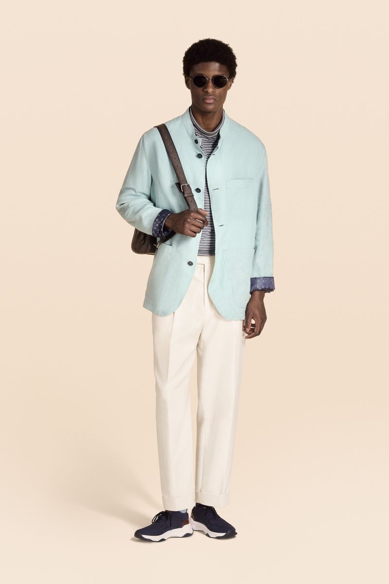 A full shot of a Black male model posing in a light-blue linen jacket, white pants, navy sneakers, and sunglasses against a beige backdrop.