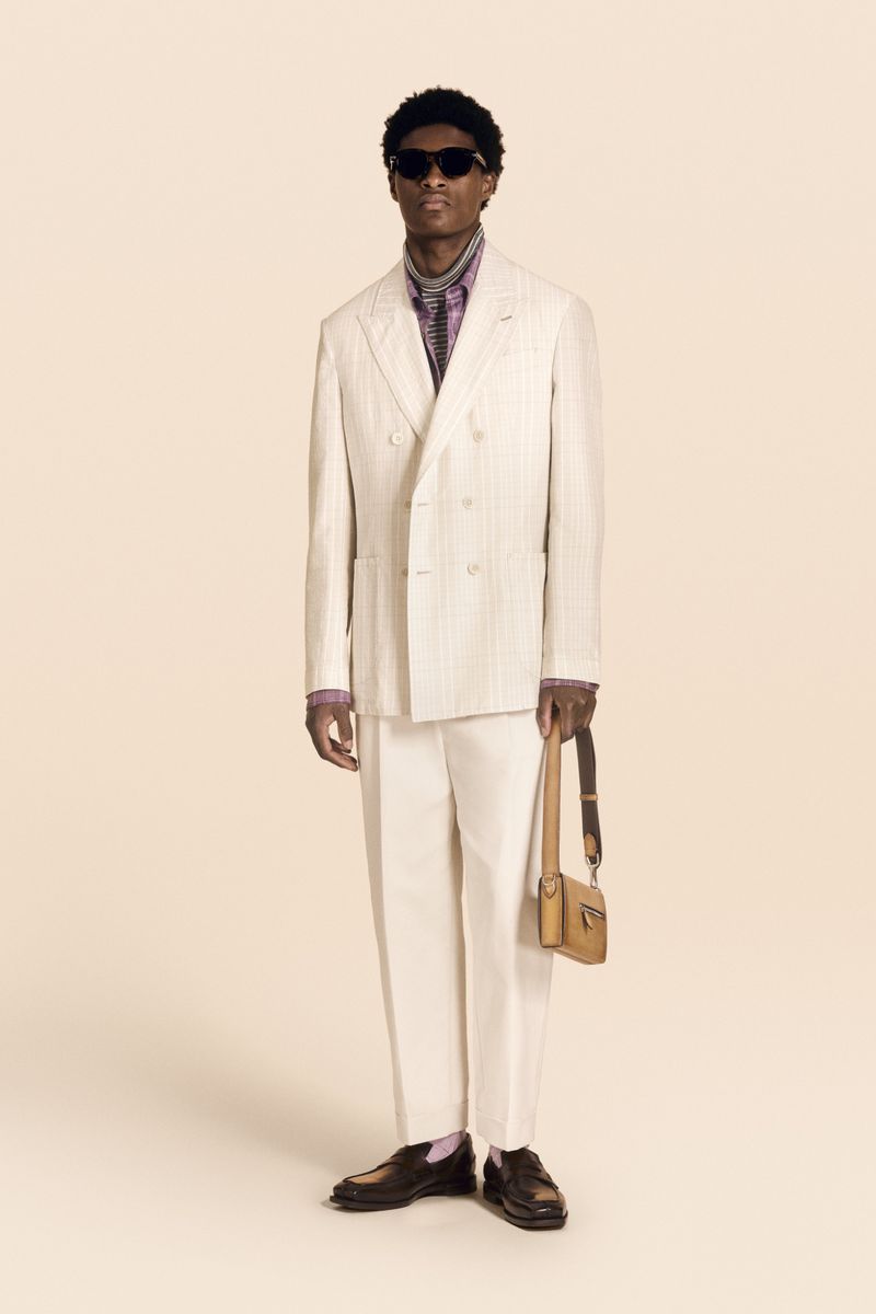 Full-length shot of an African American adult male model in a light-colored suit, holding a handbag, posing in a bright photo studio.