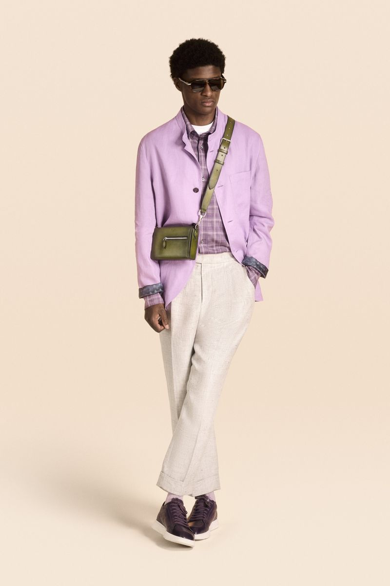 A stylish African-American man poses in a lilac jacket, white pants, olive-green bag, and sunglasses against a neutral backdrop.