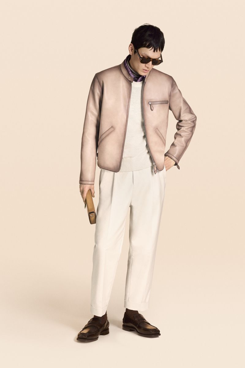 An adult male stands indoors in a tan leather jacket, white pants, brown shoes and sunglasses, holding a brown leather bag.
