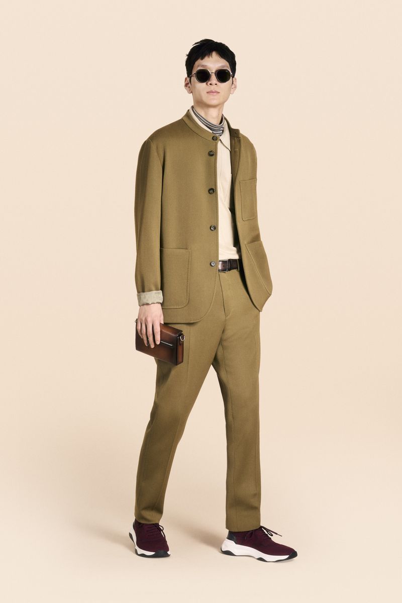 A full shot of a male model in a brown suit with sunglasses, holding a clutch, against a light peach background in a fashion editorial.