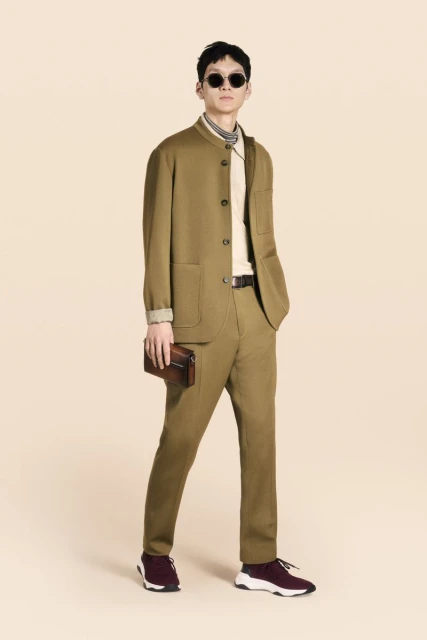A full shot of a male model in a brown suit with sunglasses, holding a clutch, against a light peach background in a fashion editorial.