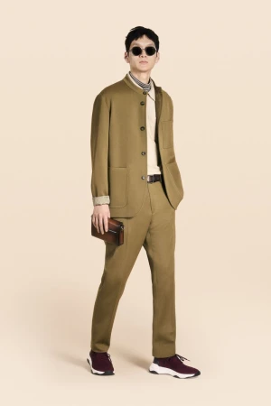 A full shot of a male model in a brown suit with sunglasses, holding a clutch, against a light peach background in a fashion editorial.
